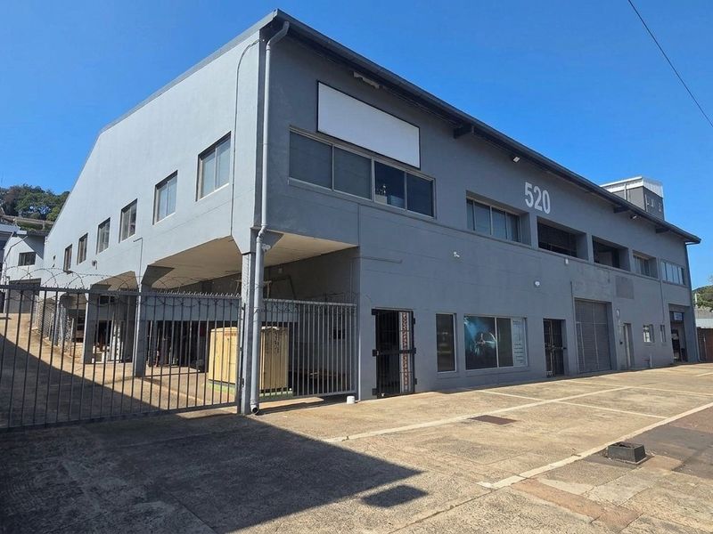 Front elevation of a multi-level industrial property featuring concrete driveway, secure perimeter fencing, and metal roller shutter access points.