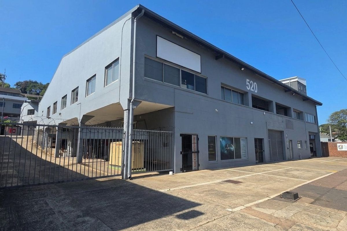 Front elevation of a multi-level industrial property featuring concrete driveway, secure perimeter fencing, and metal roller shutter access points.