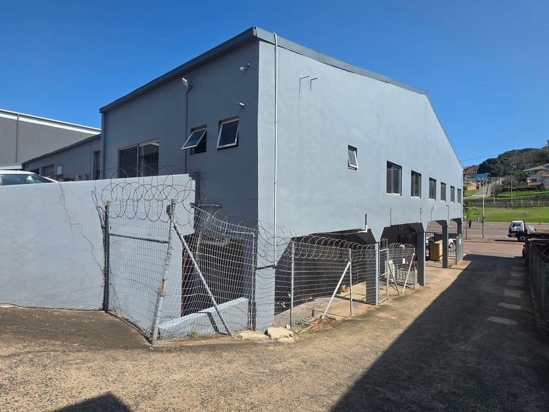 Rear exterior view of the industrial property featuring a concrete facade, security fencing with razor wire, and an asphalt driveway area.