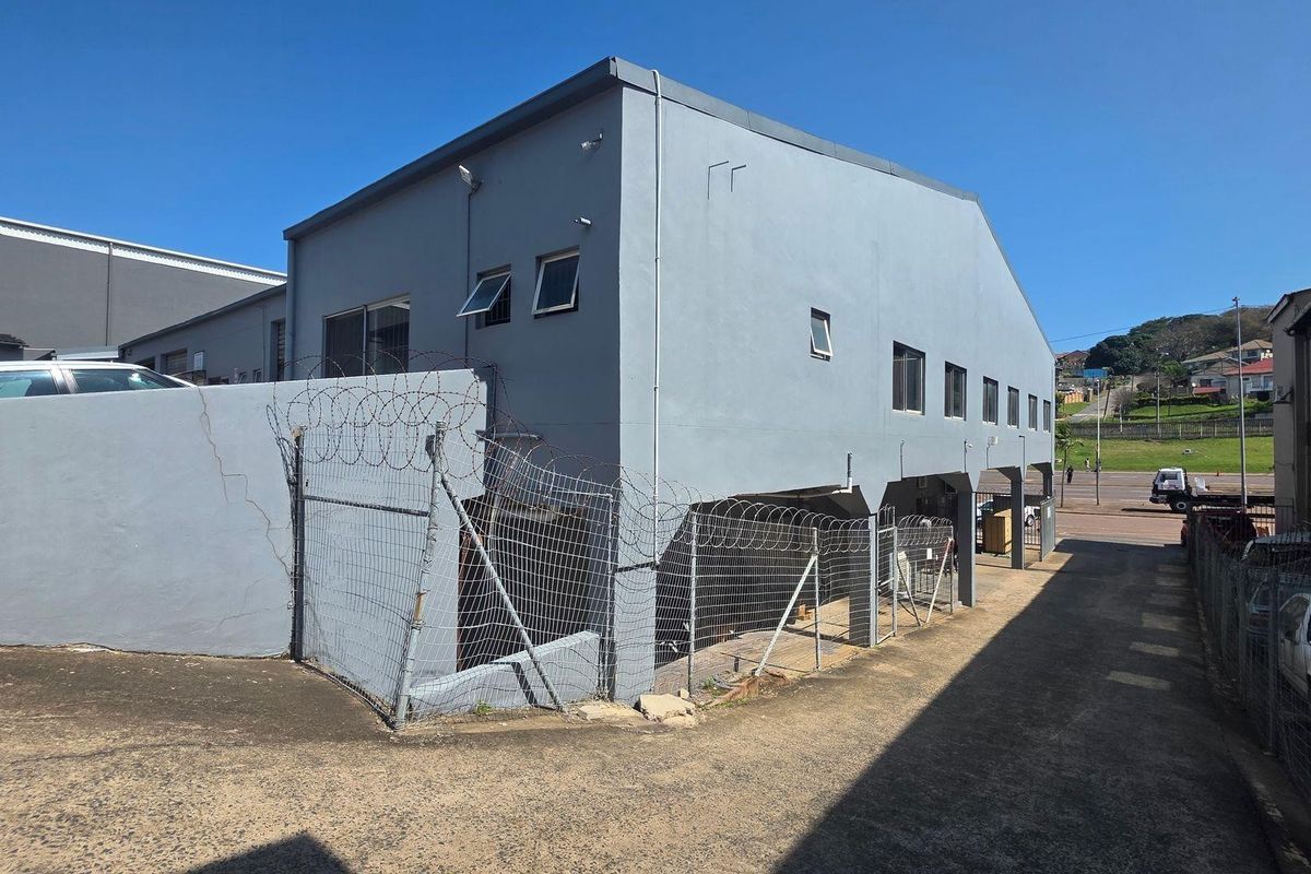 Rear exterior view of the industrial property featuring a concrete facade, security fencing with razor wire, and an asphalt driveway area.