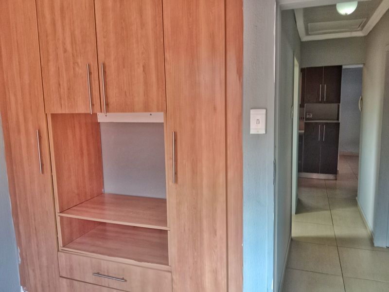 A hallway featuring built-in wooden cabinetry with integrated shelving and storage, leading through to an adjacent kitchen area with tiled flooring throughout.