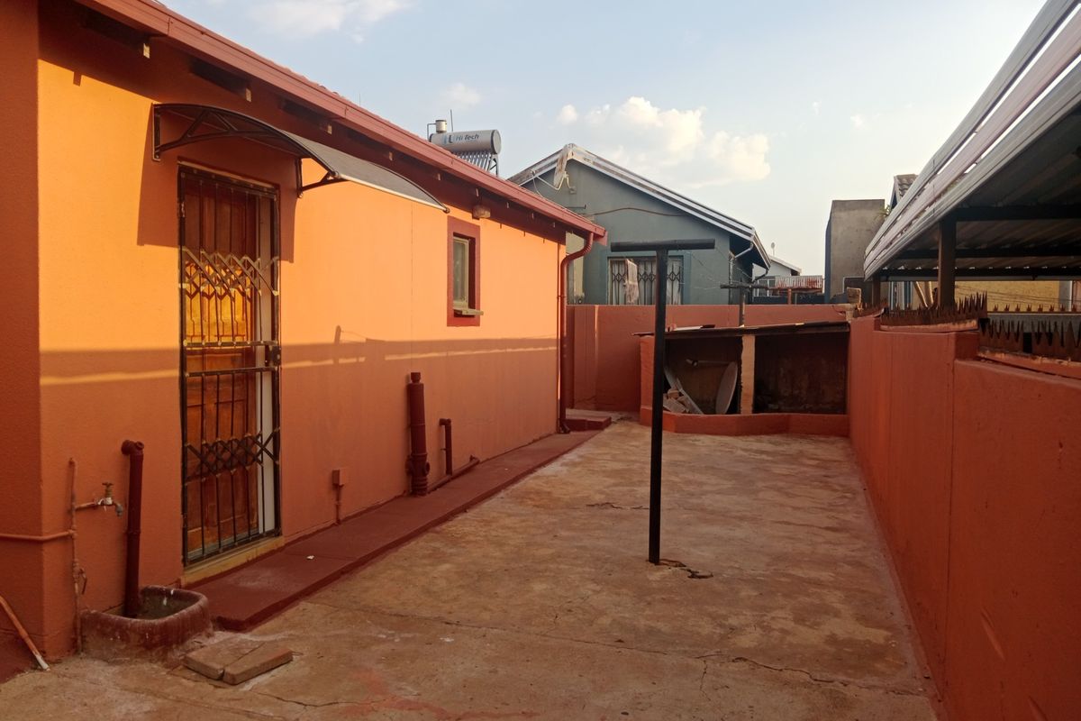 Rear exterior area featuring concrete paving, perimeter walling, and a security gate entrance, with a solar water heater visible on the roof.