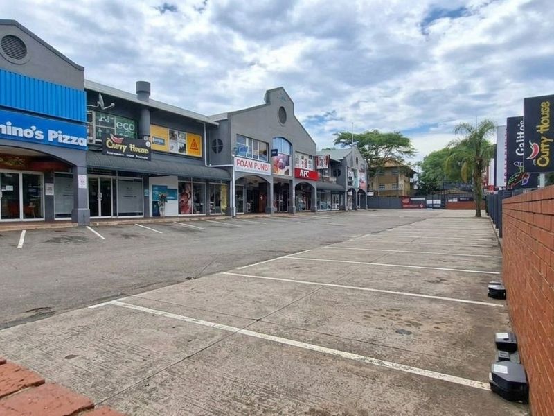 Exterior view of a commercial strip mall featuring multiple storefronts, paved parking area, and a brick boundary wall.