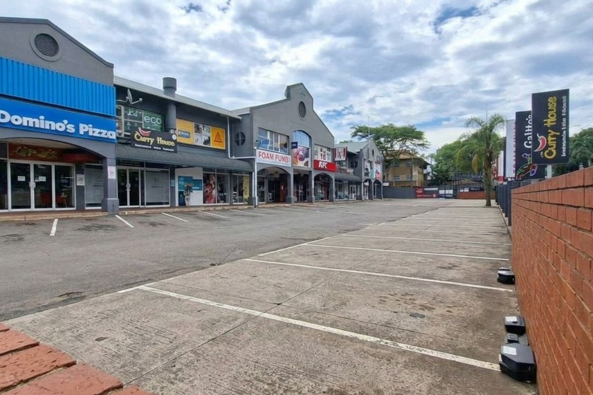Exterior view of a commercial strip mall featuring multiple storefronts, paved parking area, and a brick boundary wall.