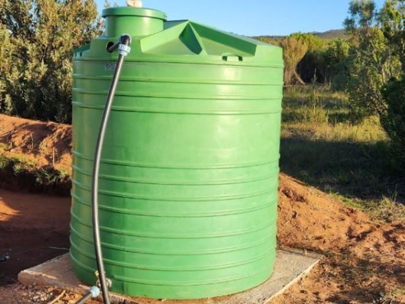 A green plastic water storage tank installed on a concrete base with associated plumbing connections, situated in an outdoor farm setting.