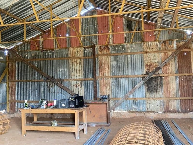 A large, rustic utility shed or workshop featuring exposed steel framing, corrugated iron walls and roof, and a natural earth floor.