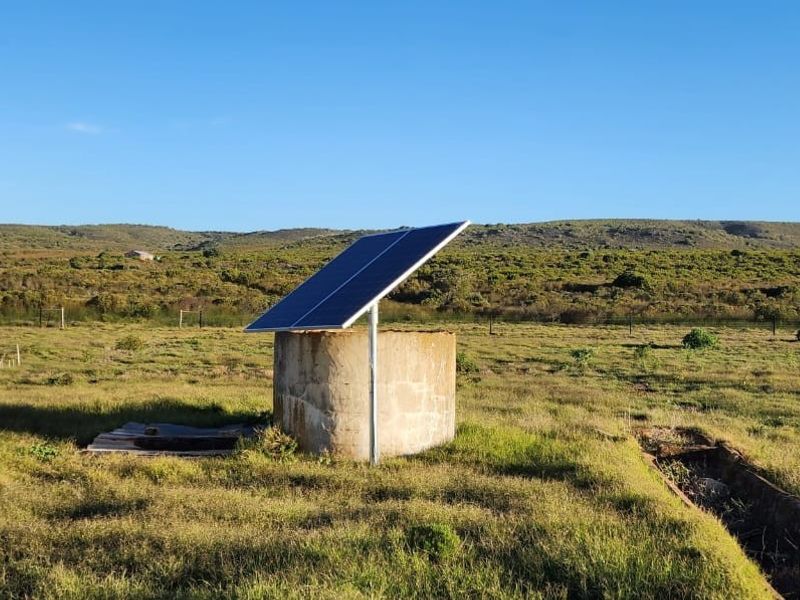 Outdoor farm area featuring a solar panel installation mounted above a concrete water reservoir and a concrete livestock trough in a rural landscape.