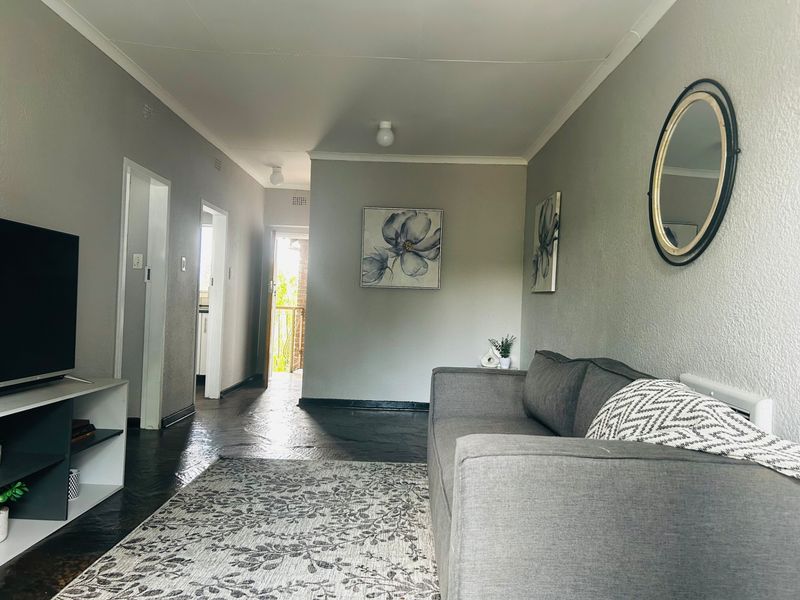 A living room featuring neutral grey walls, coved ceilings, and dark flooring, providing a functional and well-lit space with access to adjacent rooms.