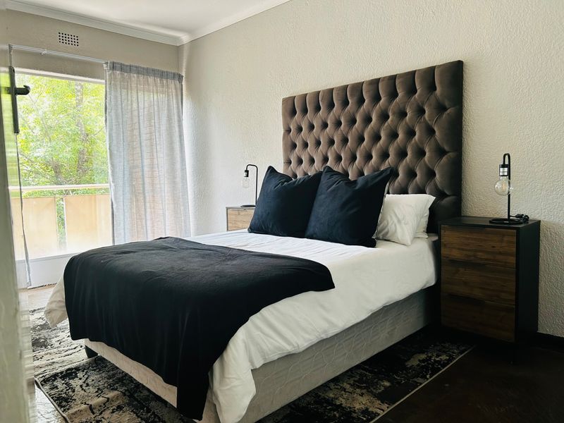 A well-maintained bedroom featuring a large tufted headboard, neutral wall finishes, and direct access to a private balcony through a sliding glass door.