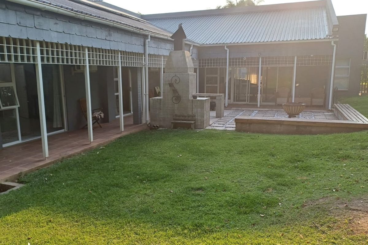 Rear exterior view featuring a covered veranda, paved patio area with a built-in braai, and a raised timber deck overlooking the garden.