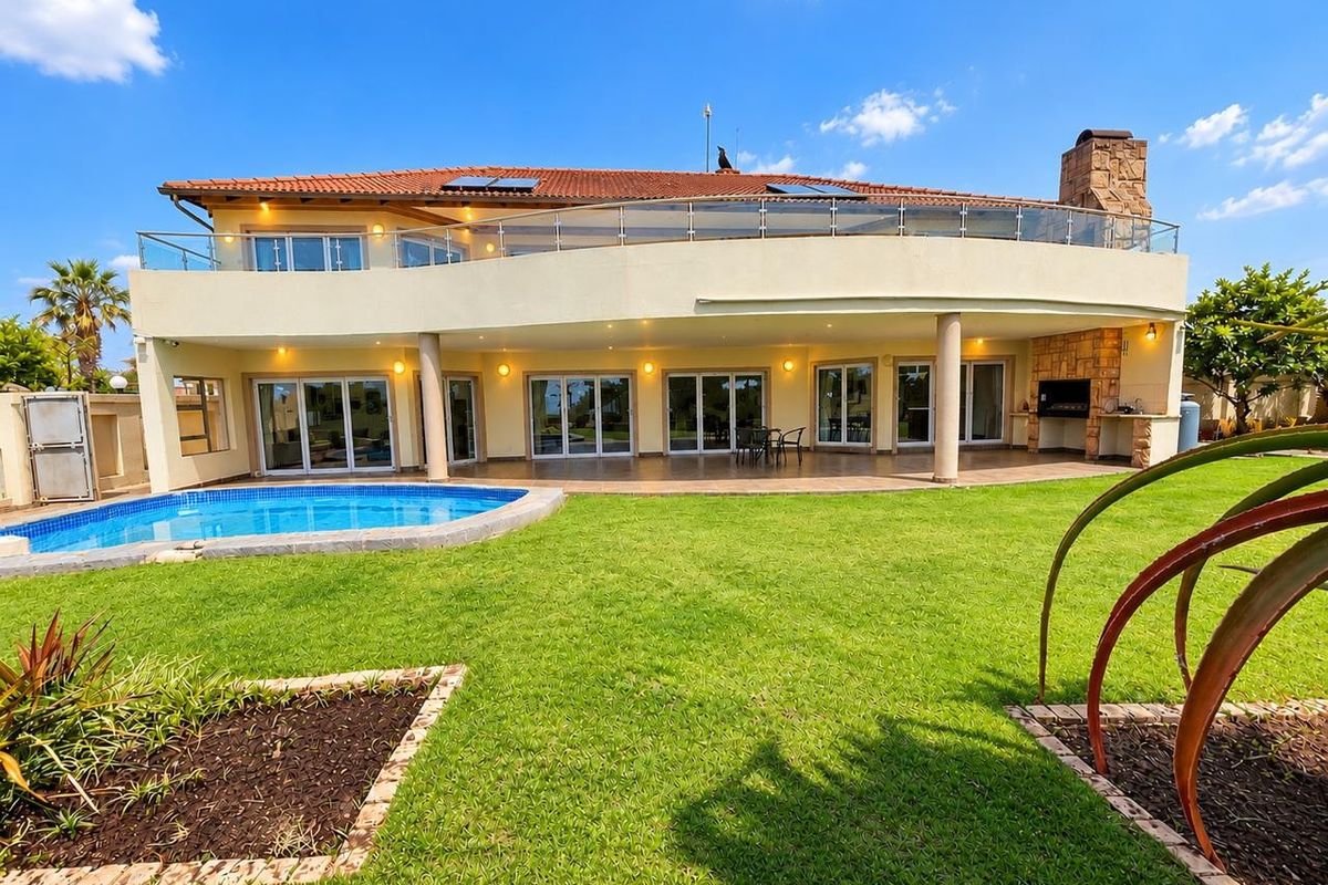 Spacious rear exterior featuring a large swimming pool, expansive covered patio with built-in braai, and an upper-level balcony with glass balustrades.