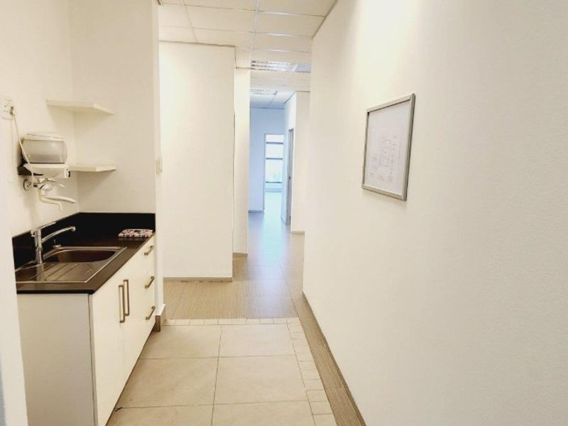A clean, functional office kitchenette area featuring a built-in sink, cabinetry, and tiled flooring, leading into a bright hallway.