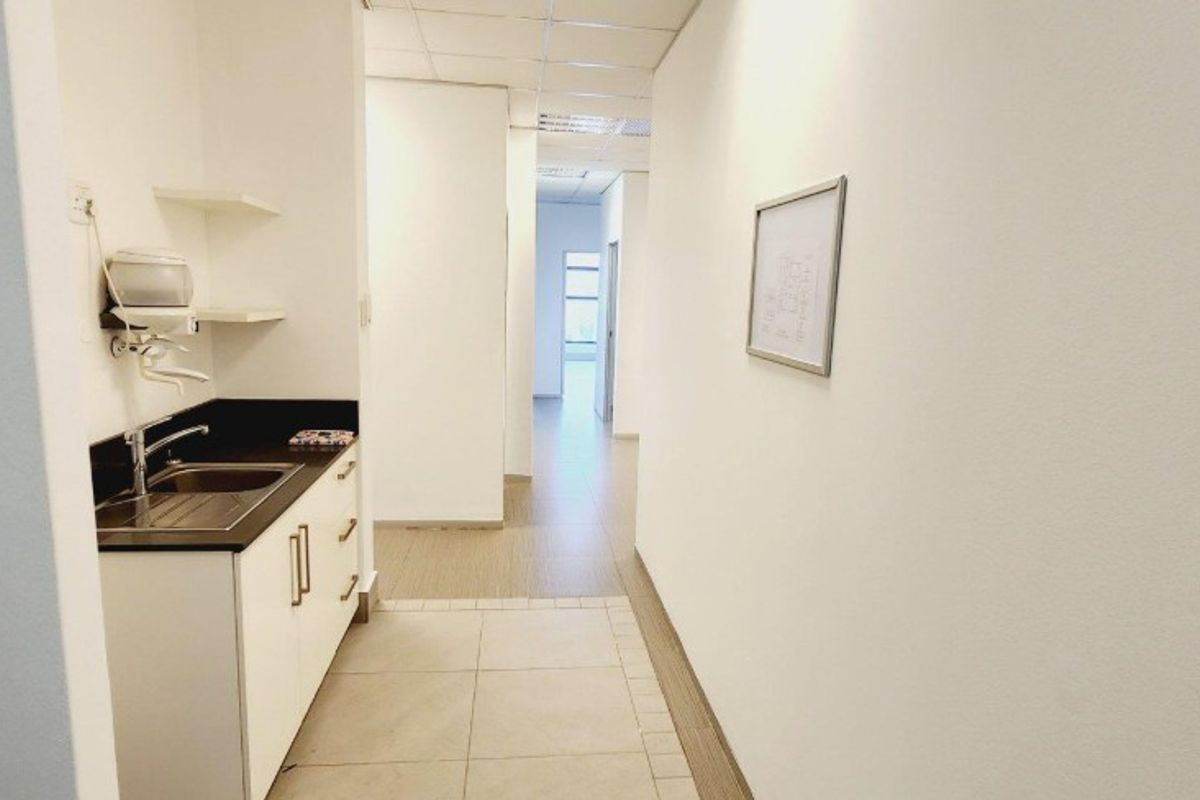 A clean, functional office kitchenette area featuring a built-in sink, cabinetry, and tiled flooring, leading into a bright hallway.