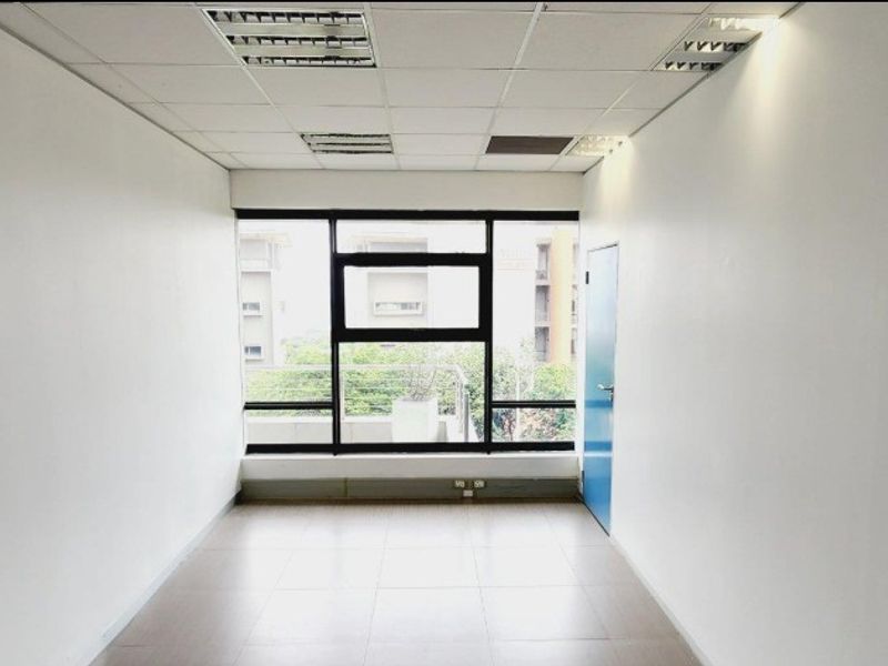 A vacant, well-lit office space featuring tiled flooring, a suspended ceiling with recessed lighting, and a large window providing natural light.