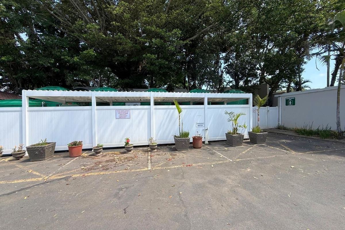 An outdoor parking area featuring a white vinyl privacy fence concealing water storage tanks, set against a backdrop of mature trees.