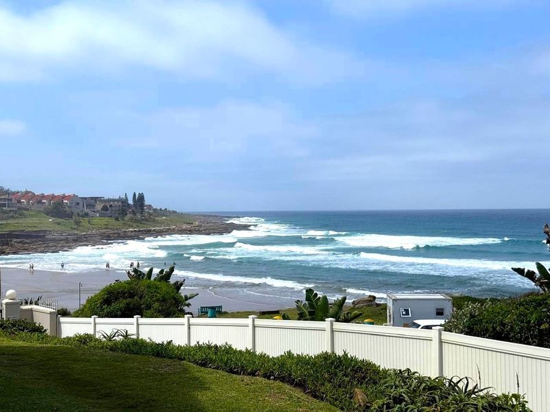 A scenic coastal view overlooking the ocean and beach, framed by a well-maintained white perimeter fence and landscaped garden area.