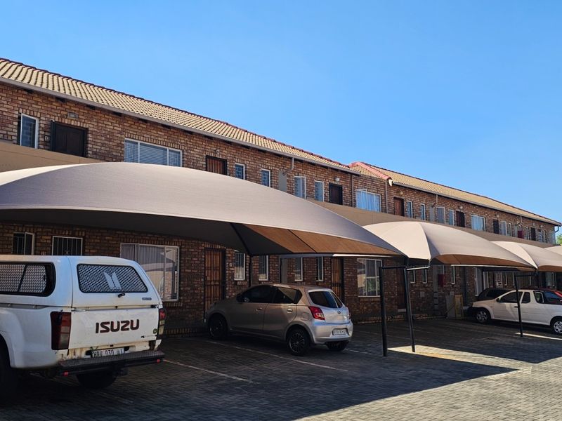 Front exterior view of a townhouse complex featuring a paved driveway with multiple shade cloth carports and brick building facade.