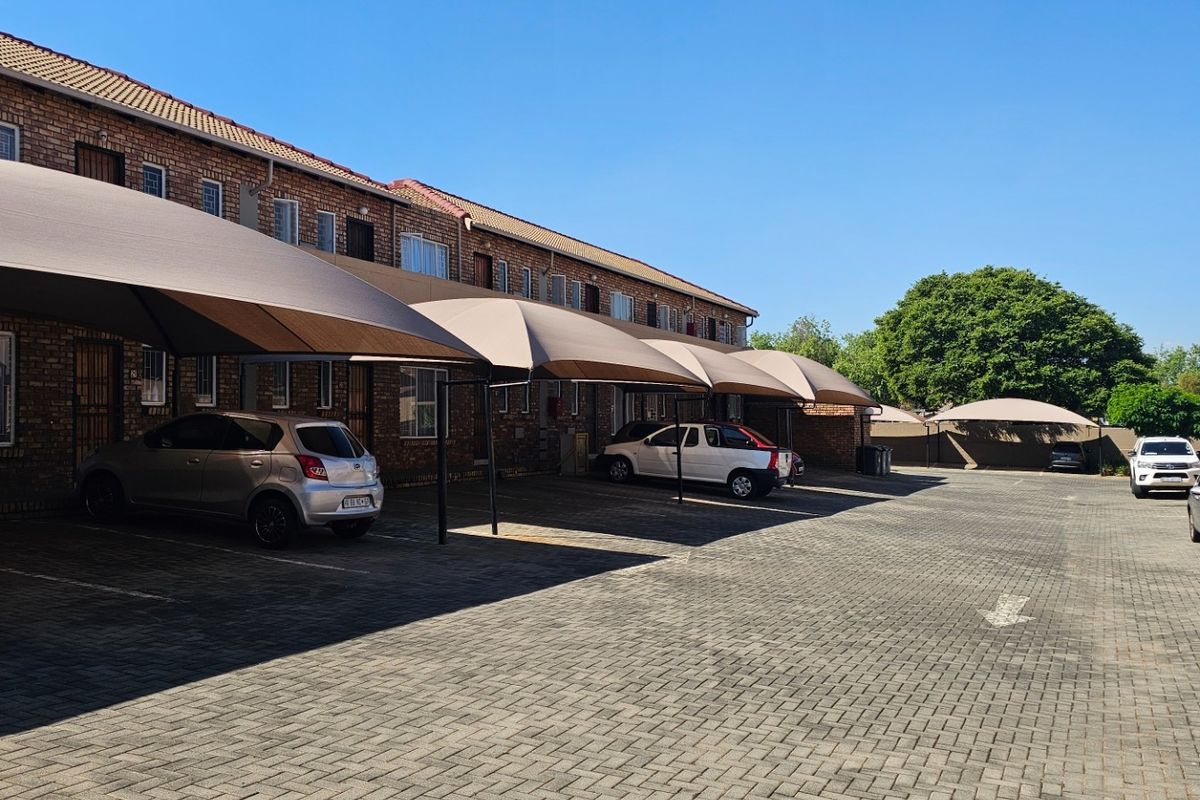 Exterior view of a townhouse complex featuring a paved parking area with multiple shade cloth carports and brick building facades.