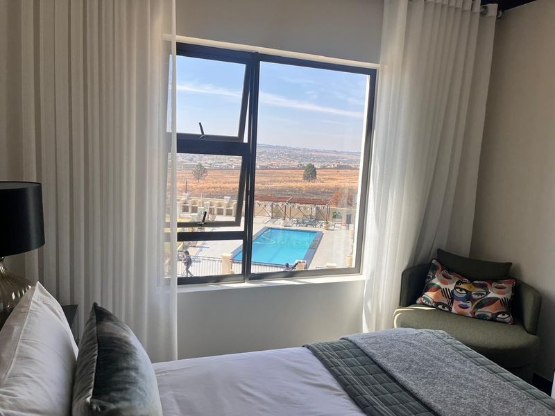 A well-lit bedroom featuring a large window with views overlooking the complex swimming pool and surrounding landscape.