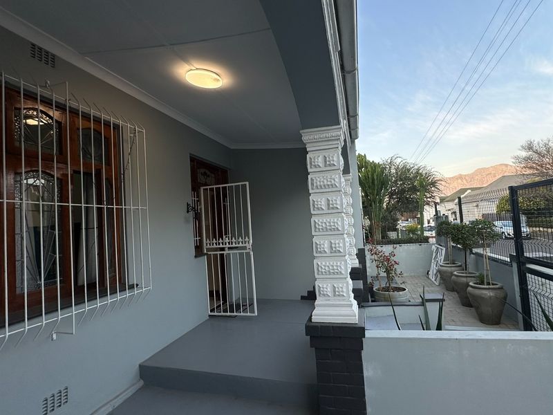 A covered front patio area featuring decorative columns, security bars on windows, and a paved entrance space with a view of the street.