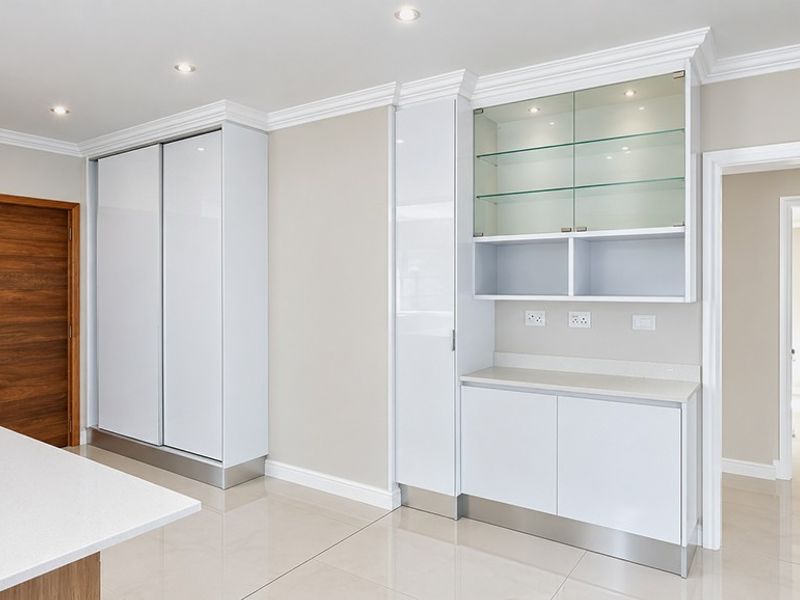 Modern kitchen area featuring sleek white built-in cabinetry, stone countertops, glass display shelving, and polished tiled flooring with recessed lighting.