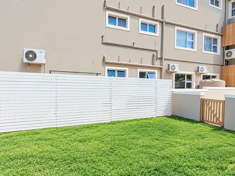 A well-maintained rear garden area featuring a private lawn, white timber slat fencing, and external air conditioning units attached to the townhouse facade.