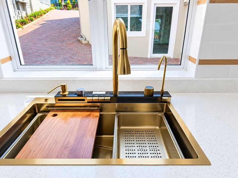 A modern kitchen sink area featuring a premium gold-tone workstation sink, gooseneck faucet, and clean quartz countertops with a subway tile backsplash.