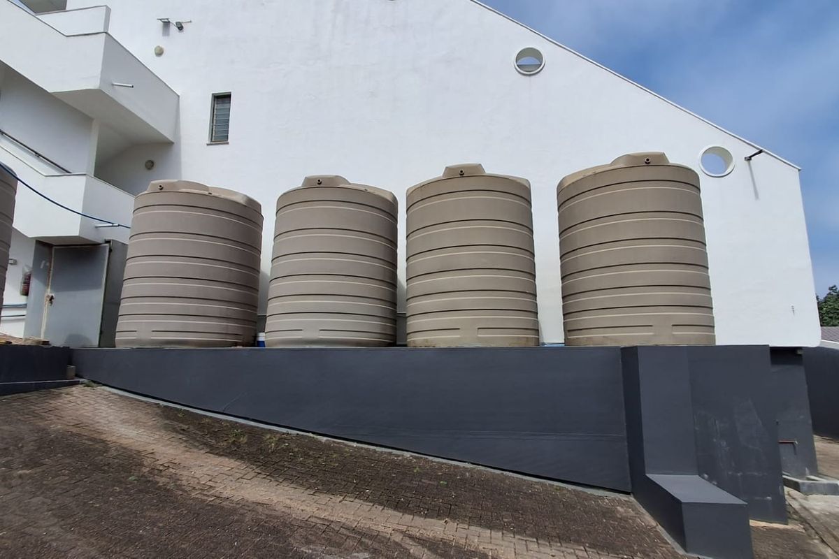 Exterior view of the property featuring a row of large water storage tanks positioned against a white building facade and a paved driveway.