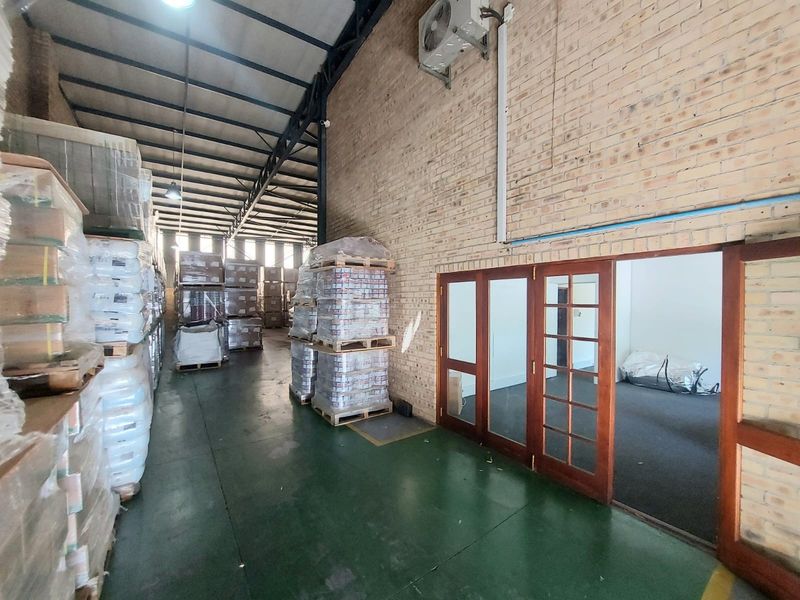 Industrial warehouse space featuring exposed brick walls, steel roof trusses, and a painted concrete floor, with access to an adjoining office area.