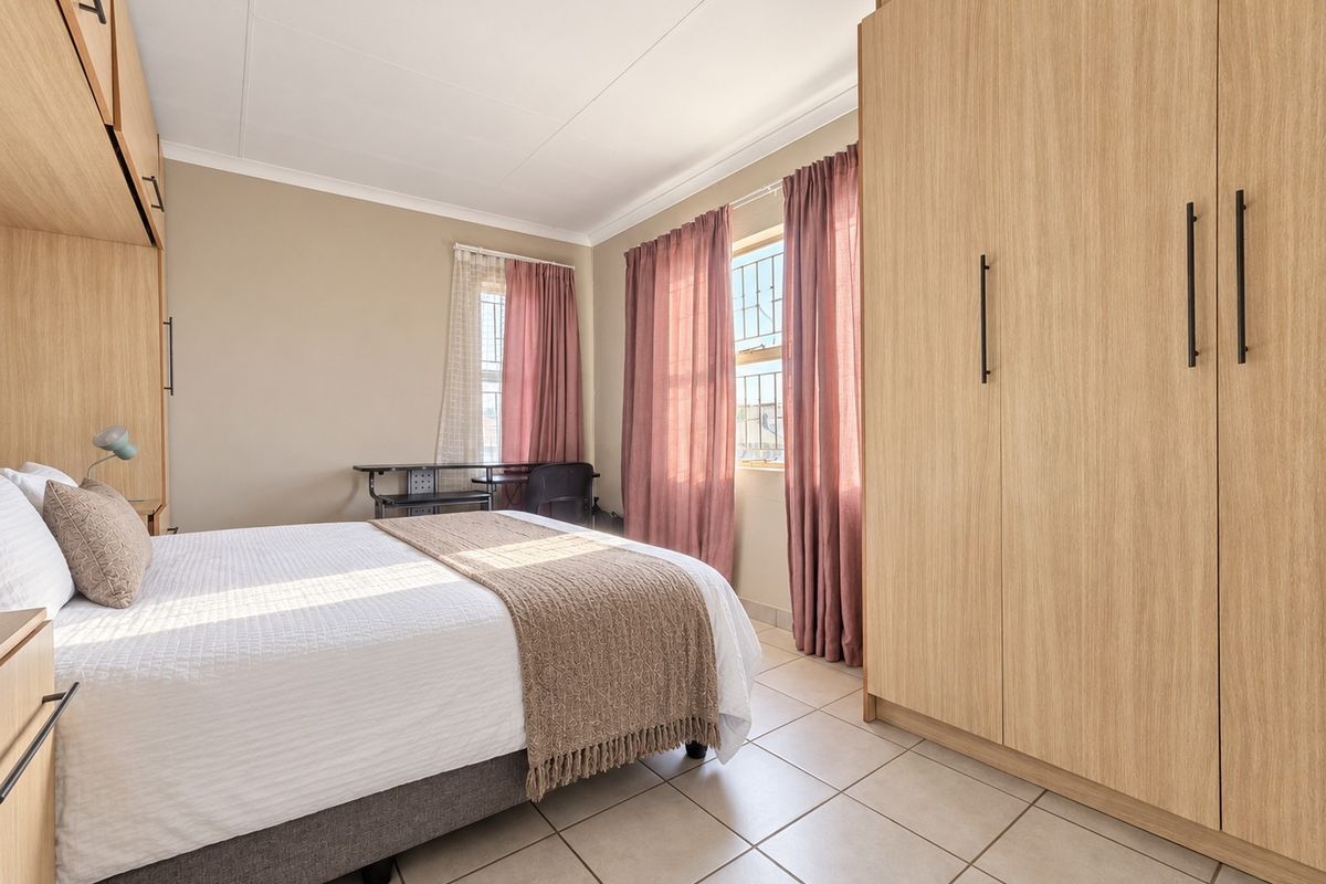 A well-maintained bedroom featuring light wood-finish built-in cupboards, neutral tiled flooring, and windows fitted with security bars.