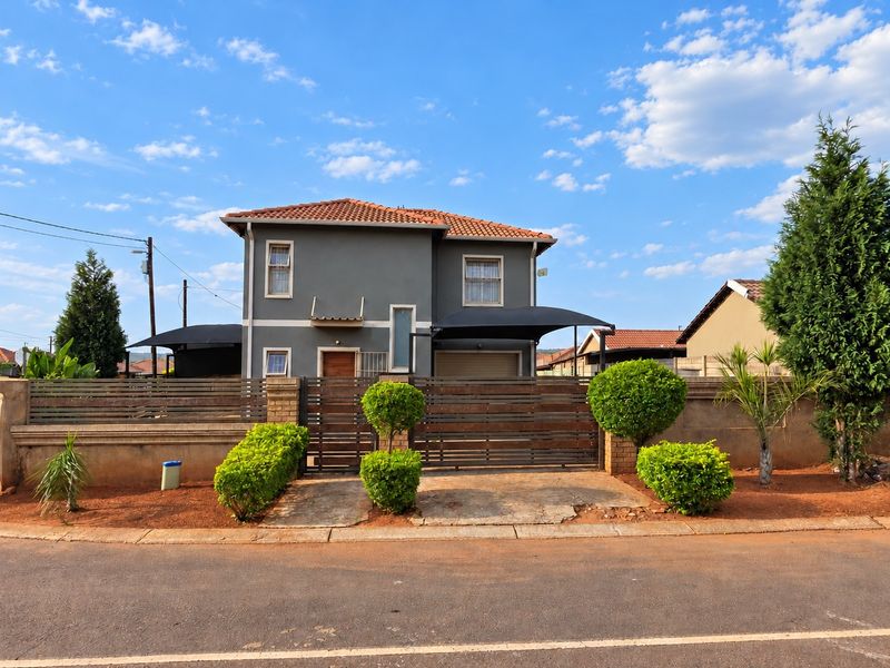 A well-maintained two-story residential property featuring a tiled roof, secure perimeter wall with gate, and a shaded carport area.