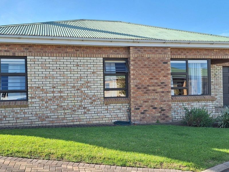 Front elevation of a face brick townhouse featuring a corrugated metal roof, aluminium window frames, and a well-maintained front lawn and paved driveway.