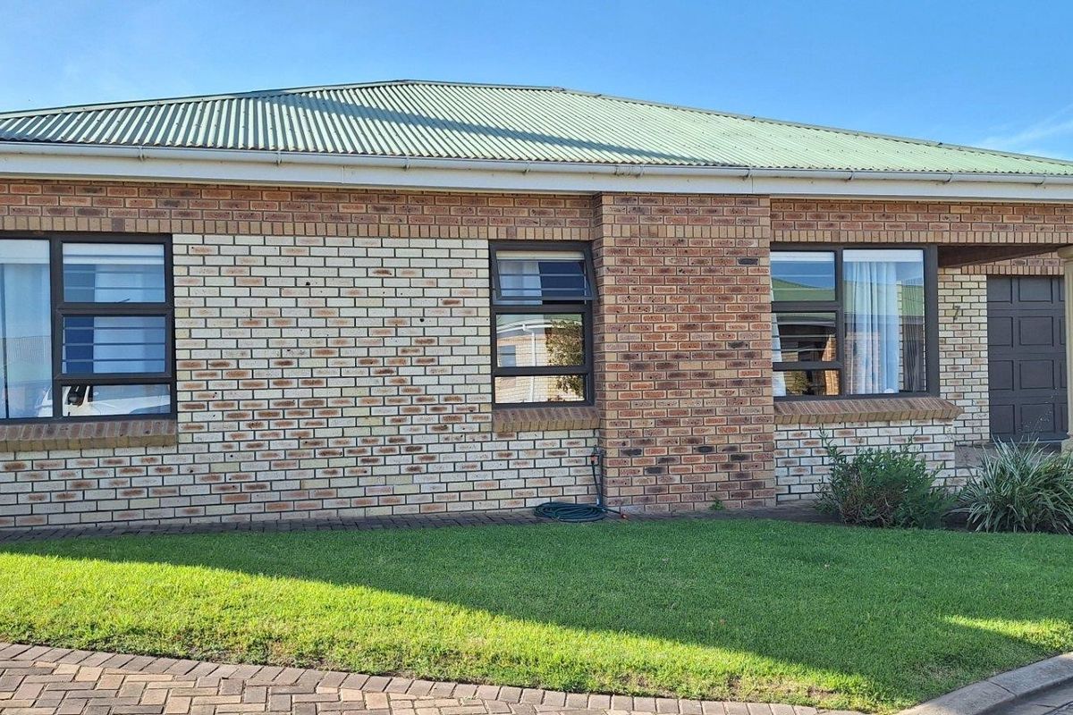 Front elevation of a face brick townhouse featuring a corrugated metal roof, aluminium window frames, and a well-maintained front lawn and paved driveway.