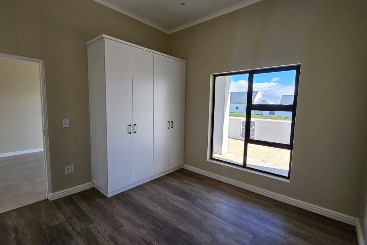 A bright, modern bedroom featuring neutral walls, dark laminate flooring, and built-in white cupboards, offering a clean and versatile living space.