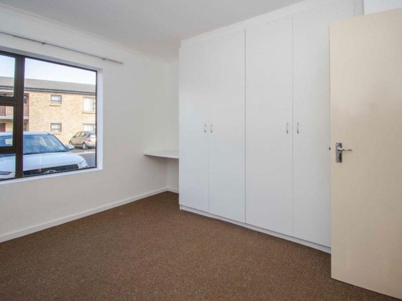 A bright, empty bedroom featuring neutral carpet flooring, a large built-in cupboard unit, and a built-in desk area under a window.