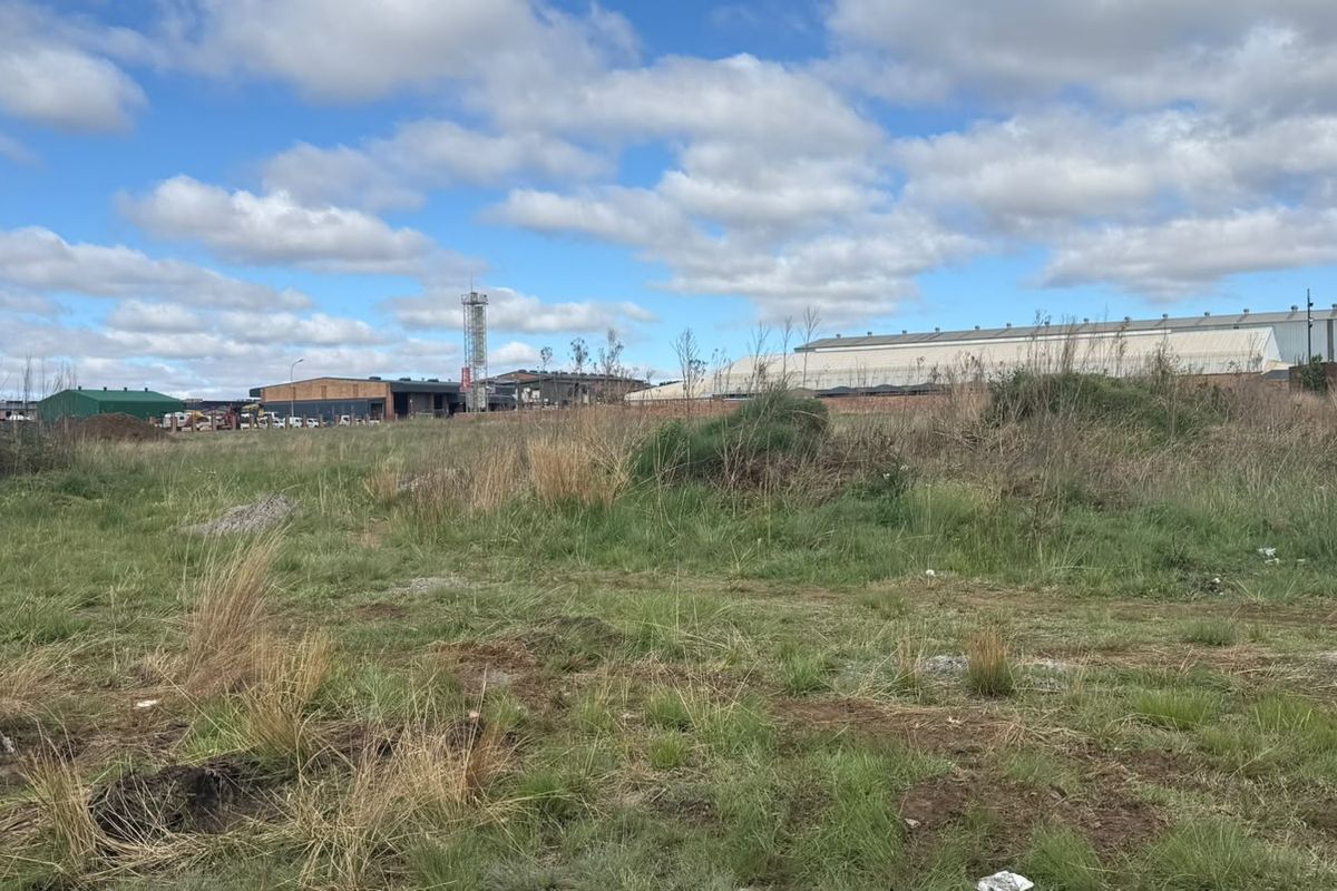 A view of an industrial site featuring warehouse buildings, a communication tower, and a perimeter wall, set against an open, undeveloped grassy field.