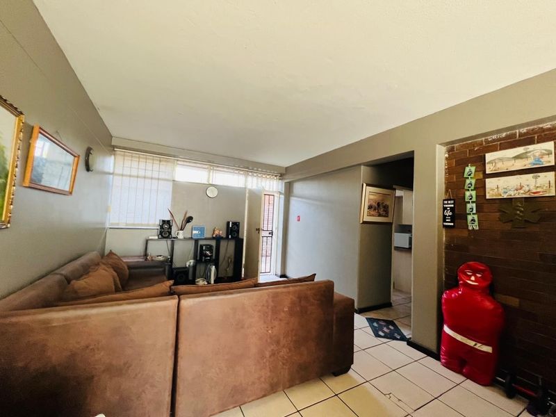 A living room featuring tiled flooring, a brick accent wall, and natural light through venetian blinds, leading to an exterior security door.