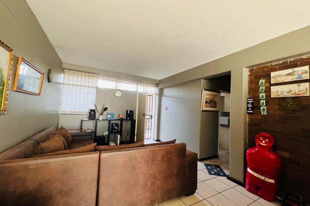 A living room featuring tiled flooring, a brick accent wall, and natural light through venetian blinds, leading to an exterior security door.