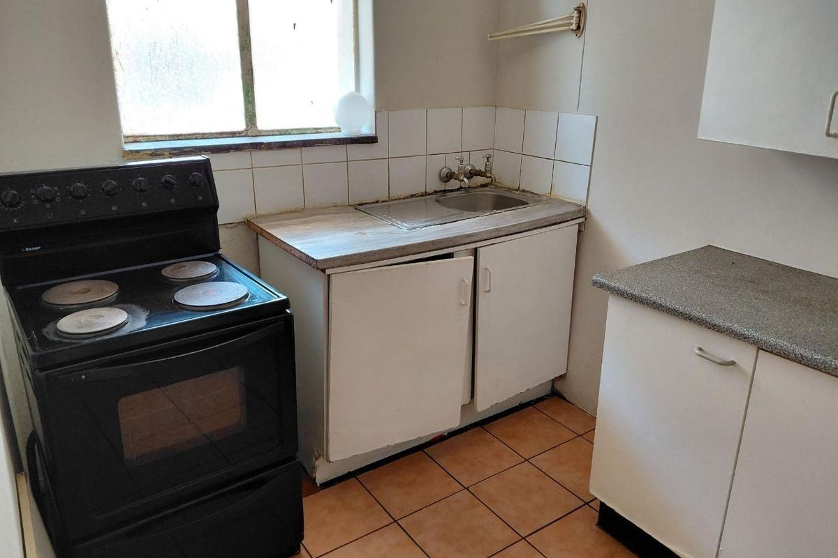 A compact kitchen featuring basic cabinetry, a stainless steel sink, tiled backsplash, and ceramic flooring, currently in need of renovation and modernization.