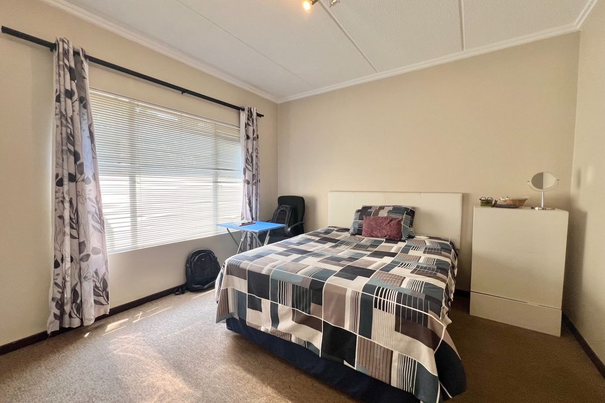 A well-lit bedroom featuring neutral wall tones, carpeted flooring, and a large window with venetian blinds and curtain rod.