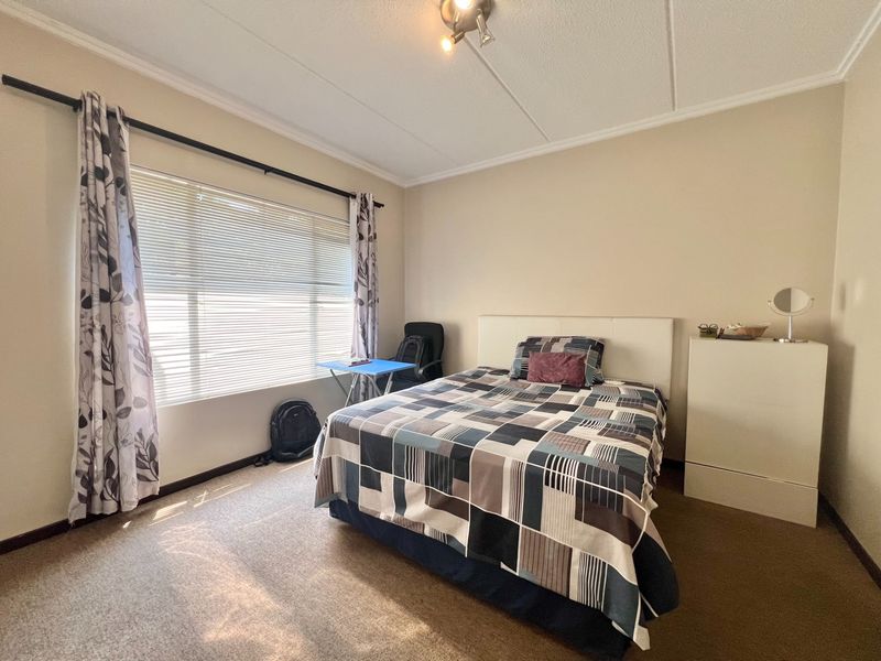 A well-lit bedroom featuring neutral wall tones, carpeted flooring, and a large window with venetian blinds and curtain rod.