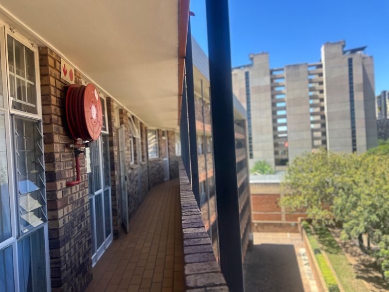 An exterior balcony walkway featuring brick masonry, tiled flooring, and a mounted fire hose reel, overlooking the apartment complex grounds.