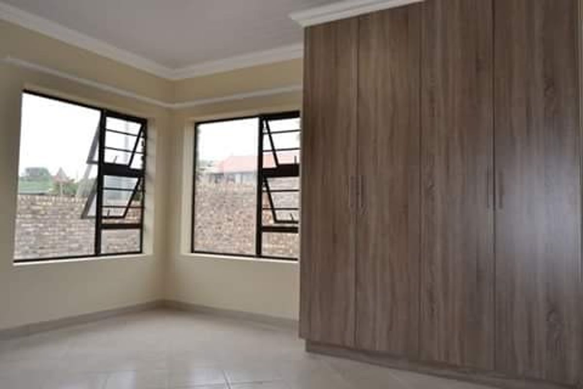 A bright, empty bedroom featuring modern wood-finish built-in cupboards, tiled flooring, and large aluminium-framed windows providing ample natural light.