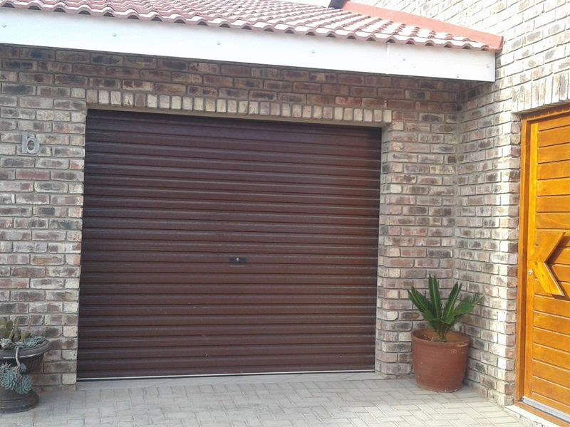 Exterior view of a brick-faced garage featuring a brown roller shutter door and a solid timber entrance door with paved driveway access.