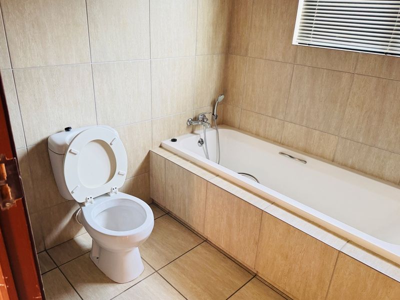 A clean, neutral-toned bathroom featuring a built-in bathtub with a handheld shower mixer, a standard toilet, and full-height wall tiling.