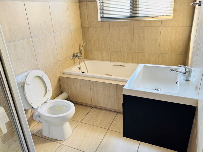 A functional bathroom featuring a built-in bathtub, vanity basin, and wall-mounted toilet with neutral floor-to-ceiling tiling.