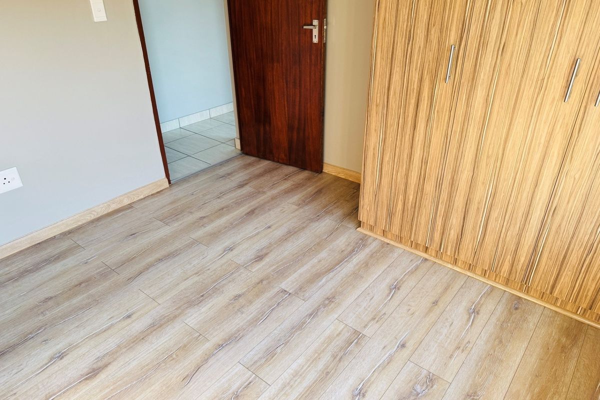 A clean, empty bedroom featuring modern laminate flooring and a built-in wooden wardrobe with a neutral wall finish.