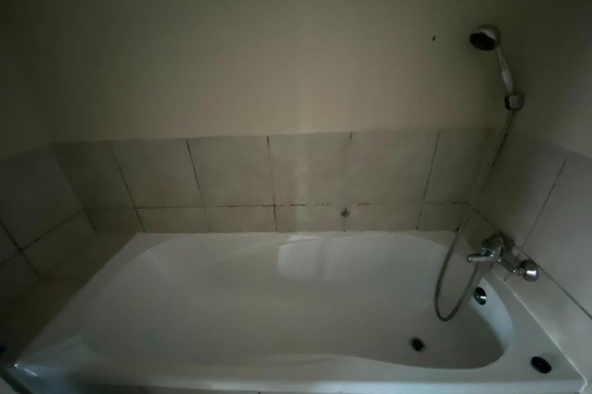 A bathroom featuring a built-in bathtub with a handheld shower fixture and tiled splashback, requiring maintenance to the grout and tile surfaces.