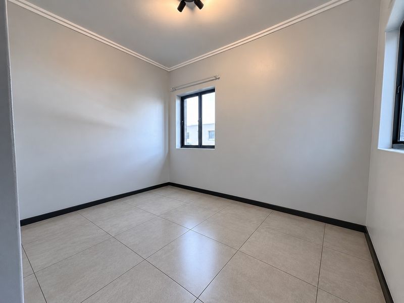 A bright, empty bedroom featuring neutral tiled flooring, clean white walls, black window frames, and decorative ceiling cornicing.
