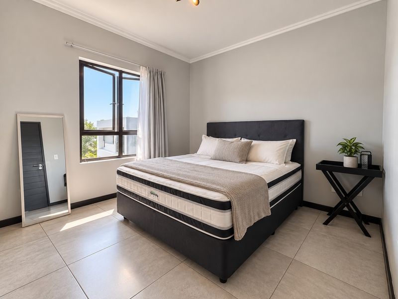 A bright and well-maintained bedroom featuring neutral tiled flooring, clean wall finishes, and a large casement window allowing for ample natural light.